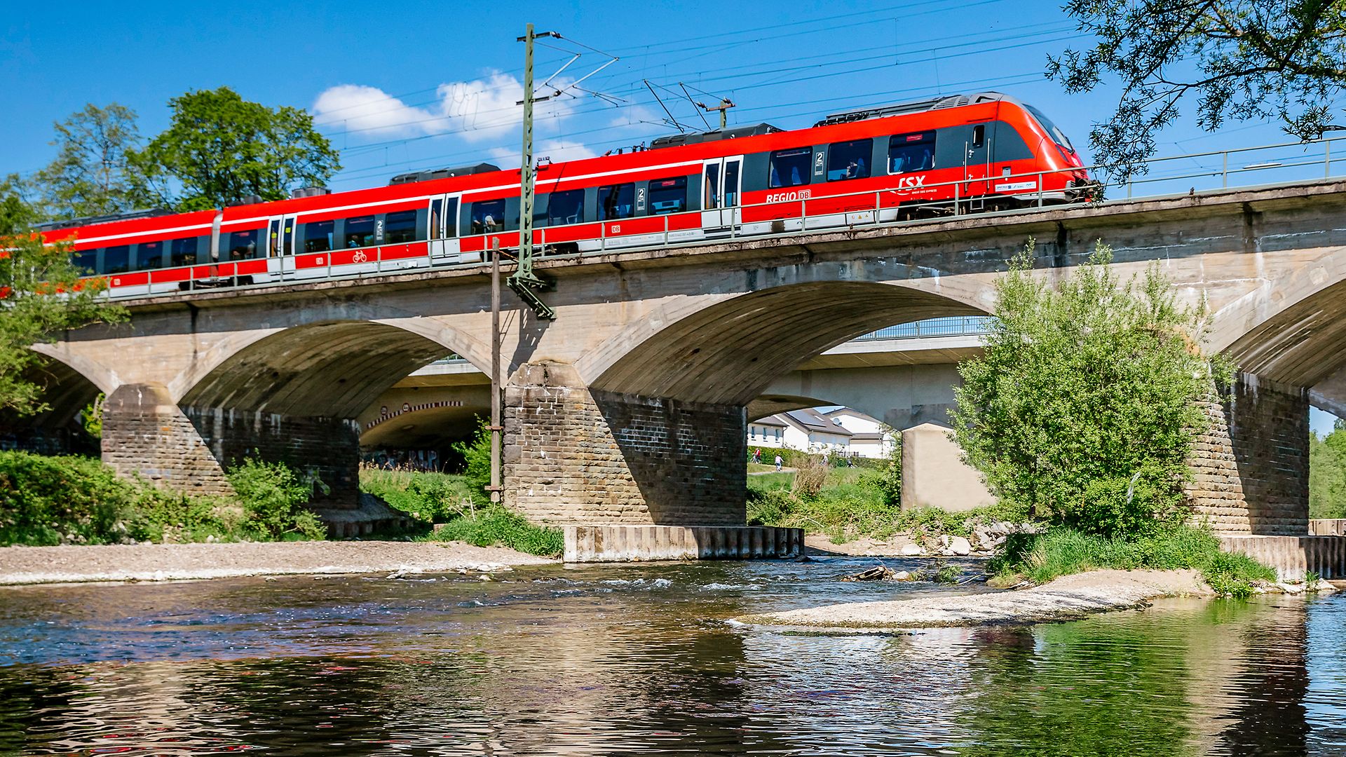 Roter RSX-Zug fährt über eine Brücke, im Vordergrund ist der Fluss sichtbar.