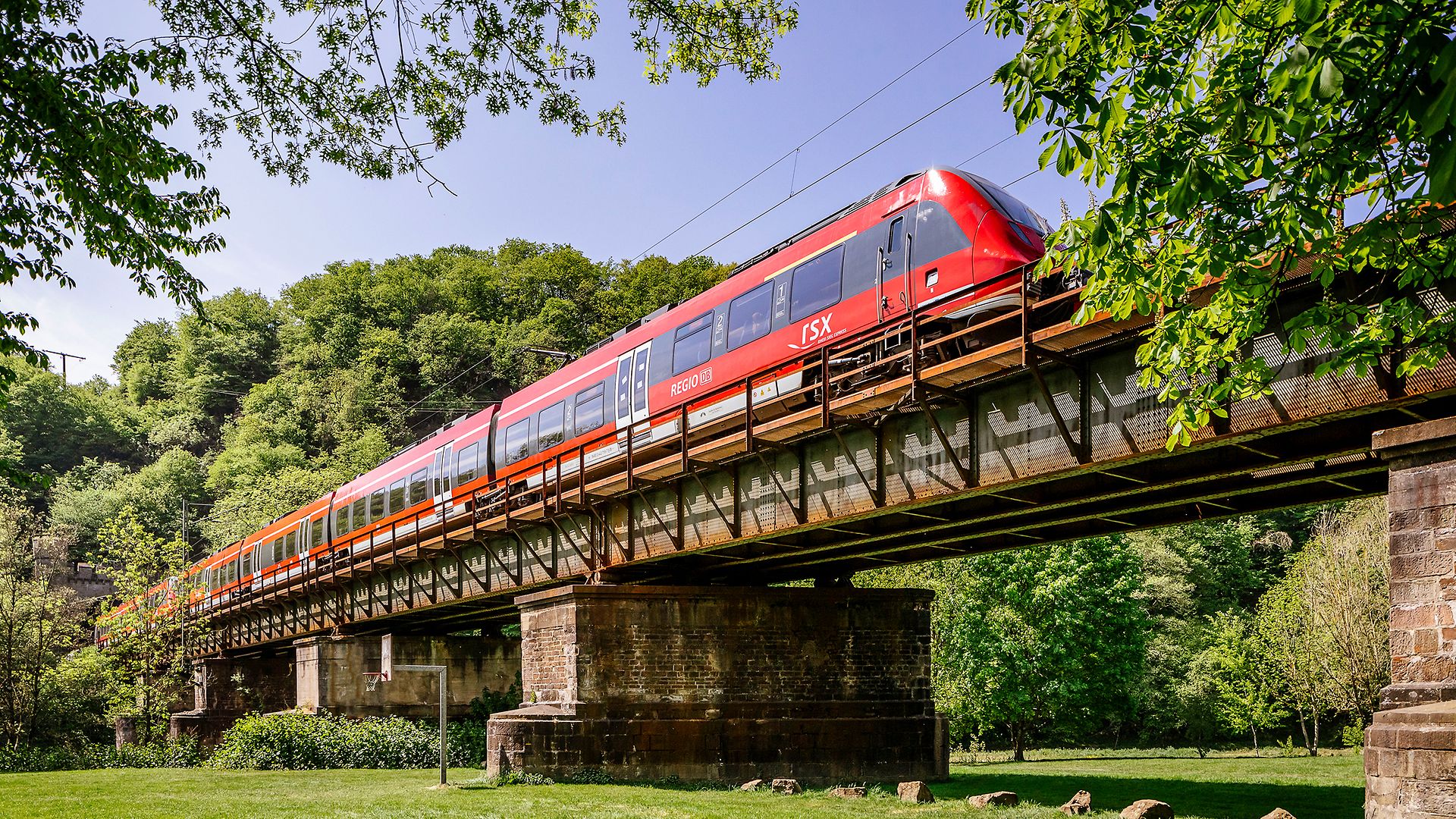 Roter RSX-Zug fährt auf einer Gleisbrücke durch eine grüne Naturlandschaft. 